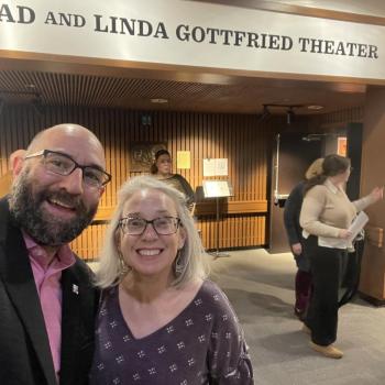 Two people smiling for a selfie inside a theater lobby with a sign reading “Brad and Linda Gottfried Theater” above them; several others are in the background.