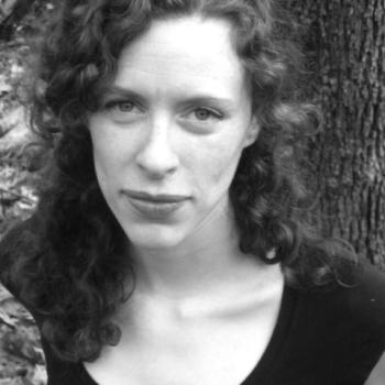 A woman with curly hair and a serious expression stands outdoors in front of a tree, wearing a dark top. The photo is in black and white.