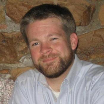 A man with a short beard and light brown hair wearing a light-colored collared shirt sits in front of a stone wall, smiling at the camera.