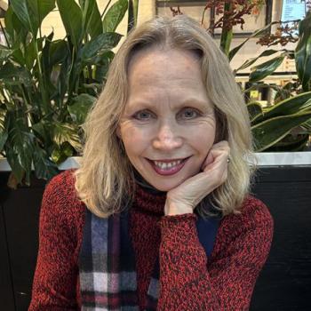 A woman with blonde hair wearing a red patterned sweater and plaid scarf sits indoors, smiling with her chin resting on her hand. Green plants and a dark partition are in the background.