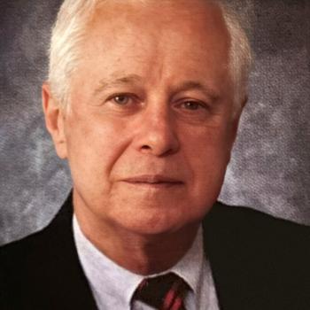 An older man with short white hair, wearing a black suit, white shirt, and red-and-black striped tie, poses against a textured gray background.