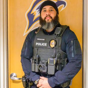 A police officer wearing a tactical vest stands in front of a wooden door with a sports team logo.