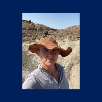 A person wearing a wide-brimmed hat, glasses, and a light shirt stands outdoors on a dusty path with rocky terrain in the background under a clear sky.