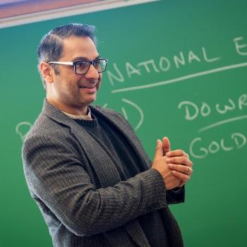 A man in glasses and a blazer stands in front of a green chalkboard with handwritten notes, gesturing as if speaking or teaching.