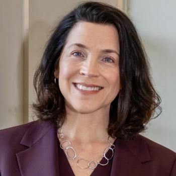A woman with shoulder-length dark hair, wearing a maroon blazer and a silver chain necklace, smiles at the camera against a neutral background.