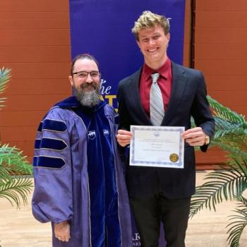 Two people stand indoors; one wears academic regalia, and the other holds a certificate and smiles. They are posing in front of a purple banner and some decorative plants.