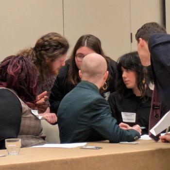 SMCM ethics bowl team  seated and standing around a table, closely discussing ethics in a conference or meeting room.