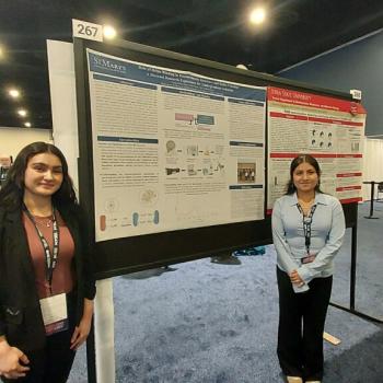 Two women stand in front of a scientific research poster at a conference, both smiling at the camera. The room is filled with other posters and attendees in the background.