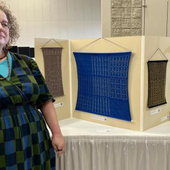 A woman stands next to a display featuring four geometric textile artworks and a tall, curved sculptural piece on a table.
