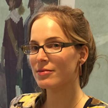 A woman with glasses and light brown hair pulled back looks at the camera. She is indoors, with a painting of two figures in historical dress in the background.
