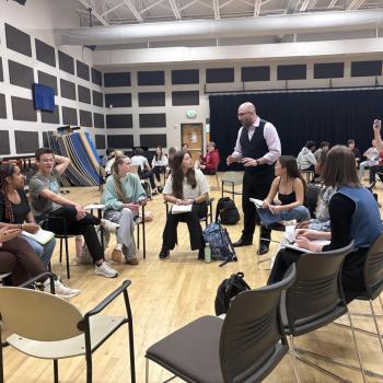 A group of students sits in a circle in a classroom while the instructor (Villada) stands and speaks; another person stands nearby and takes a photo with their phone.