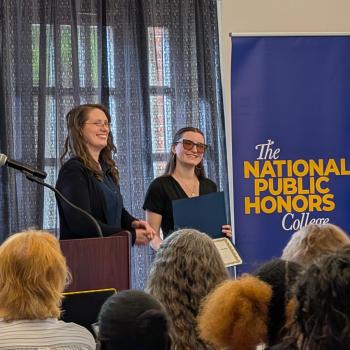 A woman receives an award on stage during a National Public Honors College event, while others applaud and a banner stands in the background.