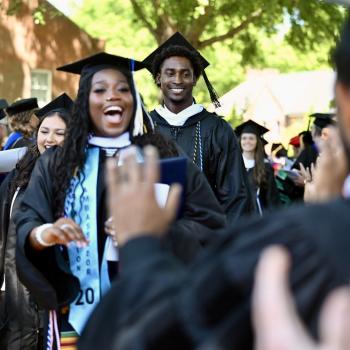 Graduates in caps and gowns walk in a procession outdoors, smiling and celebrating as people applaud around them.