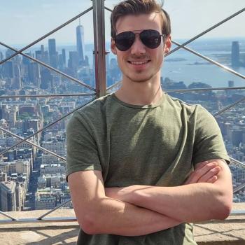 A man in sunglasses and a green shirt stands with arms crossed on an observation deck, with a city skyline and skyscrapers in the background.