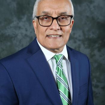 Older man with white hair and glasses, wearing a navy suit, white shirt, and green striped tie, posing in front of a mottled gray background.