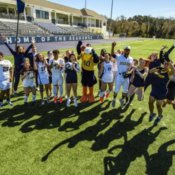 A group of athletes in sports uniforms and a mascot jump and pose for a group photo on a turf field in front of a stadium building.