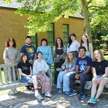 A group of eleven people pose for a photo outside on benches in front of a brick building and trees on a sunny day.
