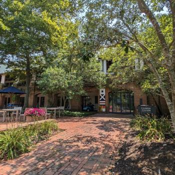 A brick walkway leads to a building entrance surrounded by trees and plants. A sign reads "Campus Center." Tables and pink flowers are visible on the left.