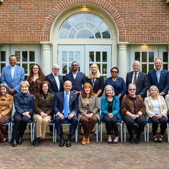 A group of 22 adults pose for a formal photo outside a brick building with arched windows, arranged in two rows—one seated and one standing.