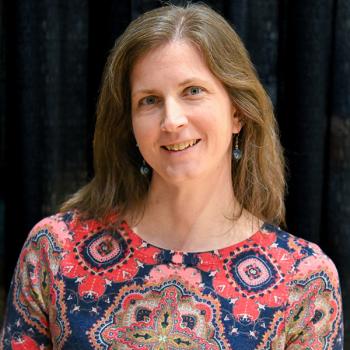 A woman with long brown hair wearing a red and blue patterned top stands in front of a black curtain, smiling at the camera.