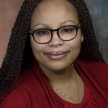 A woman with brown hair and glasses is wearing a red top, smiling at the camera against a neutral background.