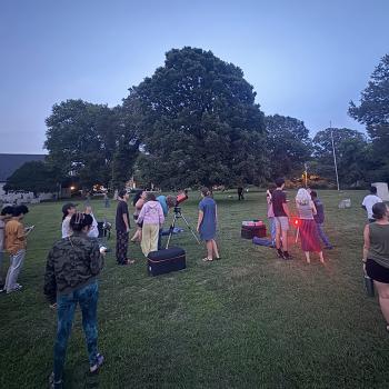 A group of people gathers on a grassy field at dusk, some standing near telescopes, while others form smaller groups. Trees and buildings are in the background.