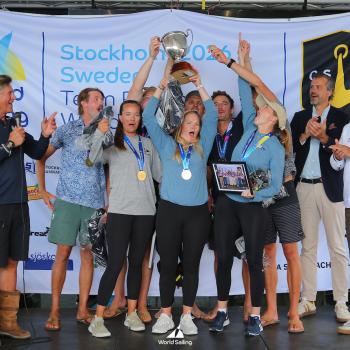 A group of people on stage celebrate with medals and a trophy at the World Sailing event, with banners and logos visible in the background. Photo by Malcom Hanes.