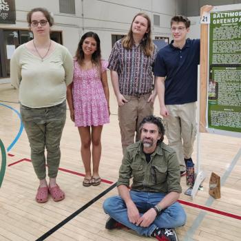 Five people pose by a science poster presentation in a gymnasium; four stand while one sits on the floor in front of the group.