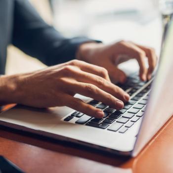 A person in business attire types on a laptop at a wooden desk, with part of a smartphone visible on the left side of the image.