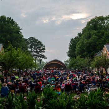 A large crowd sitting in lawn chairs facing an outdoor amphitheater stage.