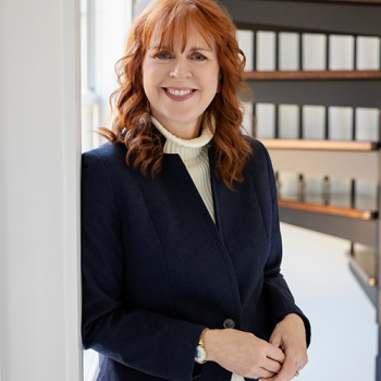A smiling woman with shoulder-length auburn hair wearing a navy blue blazer over a cream-colored turtleneck.