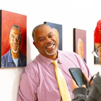 A smiling man in a pink button-down shirt and yellow tie stands in a gallery next to a portrait of a man, while a person in the foreground photographs him with a phone.