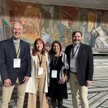 Four adults stand together outside in front of a mural wall, smiling at the camera.
