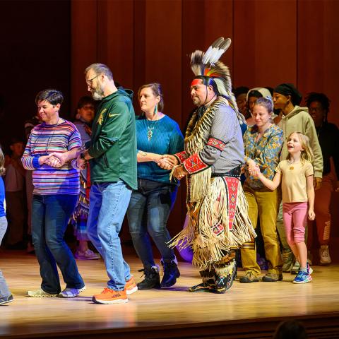 A group of people, including a man in traditional Native American attire, hold hands and form a line while dancing on a wooden stage.